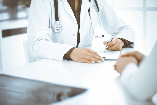 Unknown woman-doctor and female patient sitting and talking at medical examination in clinic, close-up. Therapist wearing green blouse is filling up medication history record. Medicine concept - Photo, image