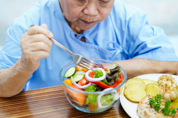 Aziatische senior of oudere oude dame vrouw patiënt eten ontbijt groente gezond voedsel met hoop en gelukkig terwijl zitten en honger op bed in het ziekenhuis. - Foto, afbeelding