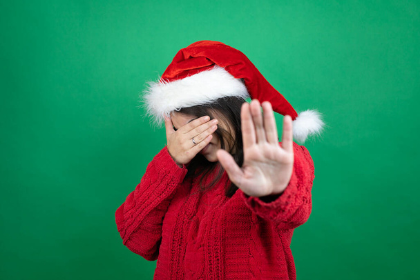 Fiatal gyönyörű nő visel karácsonyi Mikulás kalap felett elszigetelt zöld háttér borító szemek kézzel, és ezzel stop gesztus szomorú és félelem kifejezés. Zavarodott és negatív koncepció. - Fotó, kép