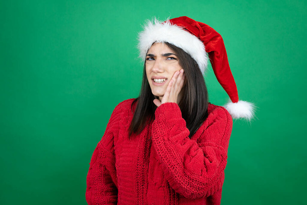 Noel Baba şapkası takan genç ve güzel bir kadın izole edilmiş yeşil arka planda diş ağrısı ya da diş ağrısı yüzünden elleriyle dudaklarına dokunuyor. - Fotoğraf, Görsel