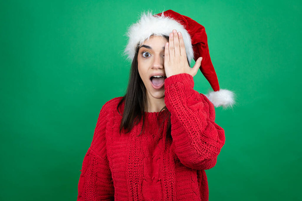 Fiatal gyönyörű nő visel karácsonyi Mikulás kalap felett elszigetelt zöld háttér borító egyik szemét kézzel, magabiztos mosoly az arcon és meglepetés érzelem. - Fotó, kép