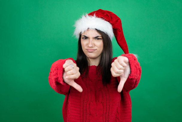 Jonge mooie vrouw dragen kerstman hoed over geïsoleerde groene achtergrond met boos gezicht, negatief teken toont afkeer met duimen naar beneden - Foto, afbeelding