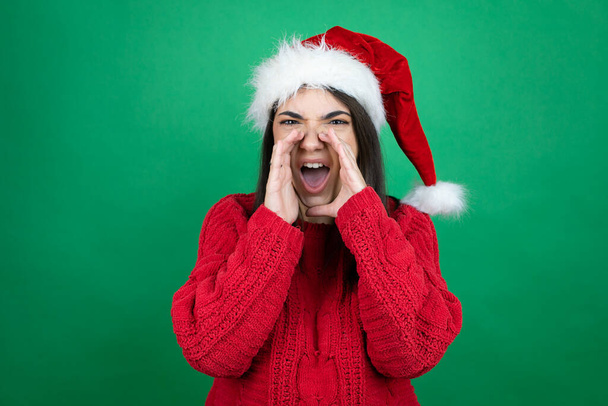 Nuori kaunis nainen yllään joulupukki hattu yli eristetty vihreä tausta huutaa ja huutaa kovaa puolelle kädet suulla - Valokuva, kuva