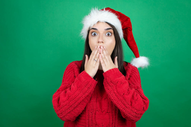 Молодая красивая женщина в рождественской шляпе Санта-Клауса на изолированном зеленом фоне с руками над ртом и удивлен - Фото, изображение
