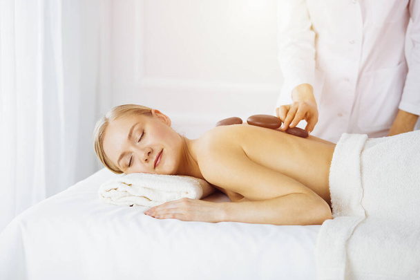 Beautiful caucasian woman enjoying warm stones procedure in sunny spa salon. Beauty concept - Foto, afbeelding