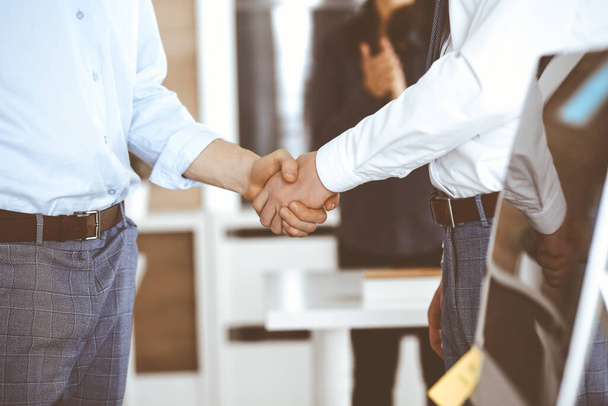 Two businessmen are shaking hands in office, close-up. Happy and excited business woman stands with raising hands at the background. Business people concept - Foto, immagini