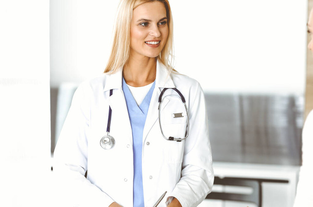 Doctor woman at work in hospital excited and happy of consulting female patient. Blonde physician controls medication history records and exam results while using clipboard. Medicine and healthcare - Foto, Imagen