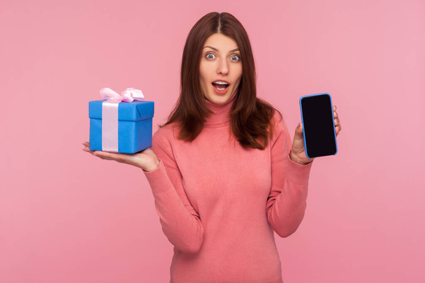 Excited brunette woman in pink sweater showing smartphone and gift with shocked amazed expression. Indoor studio shot isolated on pink background - Foto, Imagem