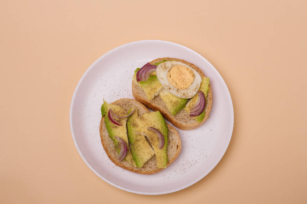 Два сэндвича с авокадо, вареные яйца и красный лук на белой тарелке на бежевом фоне. Минималистическая композиция. - Фото, изображение