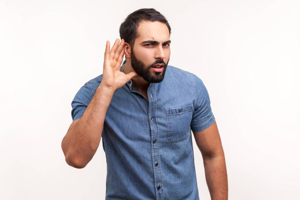 Concentrated nosy man with beard in blue shirt holding hand near ear, listening to interesting talks and private secrets, spying. Indoor studio shot isolated on white background - Valokuva, kuva