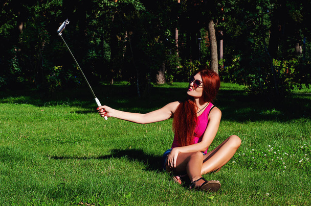 Ένα όμορφο κορίτσι με κόκκινα μακριά μαλλιά τραβάει φωτογραφίες του εαυτού της στο τηλέφωνο με τη βοήθεια ενός μονόποδα που κάθεται σε ένα πάρκο στο γρασίδι - Φωτογραφία, εικόνα