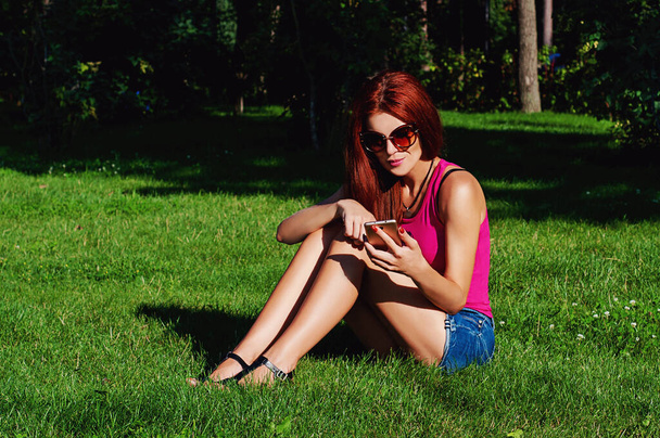 Krásná dívka s červenými dlouhými vlasy sedí na trávě v parku, má v ruce telefon. Emocionální fotografie - Fotografie, Obrázek