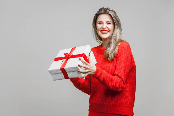 Portrait of charming blonde woman with red lipstick in bright casual sweater, holding wrapped gift box and smiling at camera, excited with surprise. indoor studio shot isolated on gray background - Photo, Image
