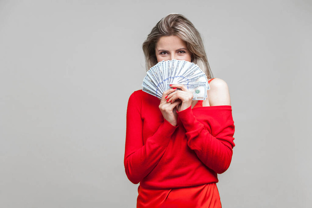 Portrait of playful rich beautiful woman in red dress standing peeking out of dollar bills with cunning eyes, hiding her half face, tricky look. indoor studio shot isolated on gray background - Foto, afbeelding