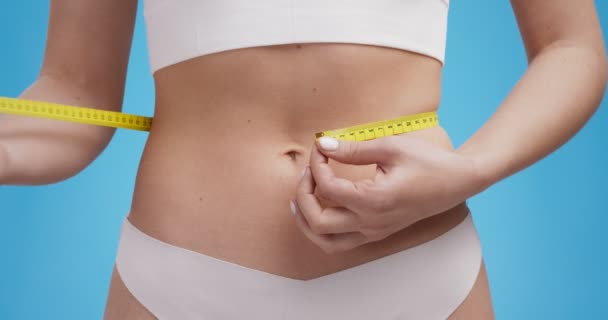 Concept de fitness et de perte de poids. Gros plan de la jeune femme mesurant sa taille avec du ruban adhésif, fond bleu - Séquence, vidéo