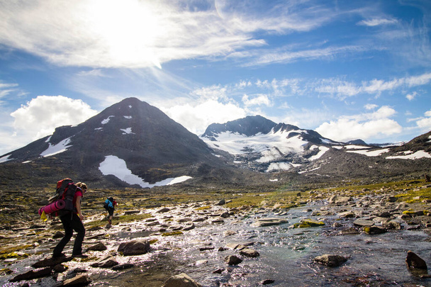 Vrouwelijke backpacker gaat door de waterstroom, met prachtige besneeuwde bergen op de achtergrond - Foto, afbeelding