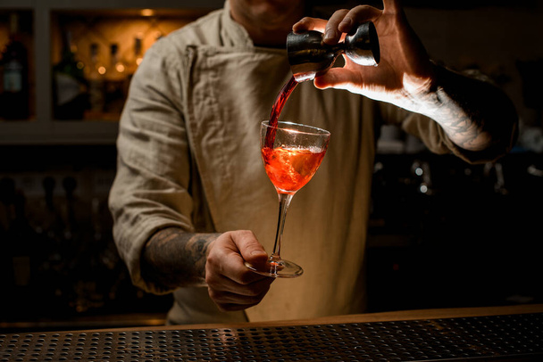 male bartender holds with hand wine glass with bright drink and adds liquid from jigger - Φωτογραφία, εικόνα