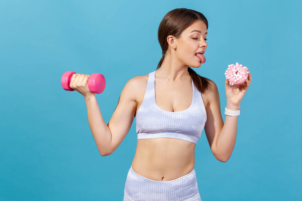 Athletic woman in white sports tights and top holding dumbbell and donut, showing tongue going to lick it, keeping diet going for sport. Indoor studio shot isolated on blue background - Zdjęcie, obraz