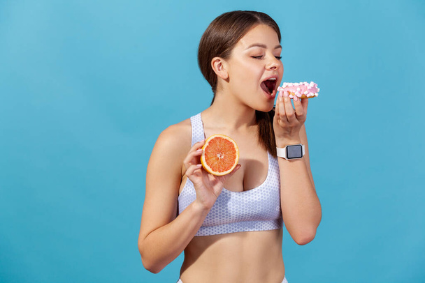 Athletic woman in sports top eating sweet junk donut instead of fresh juicy orange, choosing junk food, lack of willpower and motivation. Indoor studio shot isolated on blue background - Fotoğraf, Görsel