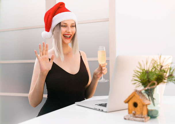 Eine fröhlich lächelnde Blondine in schwarzem Kleid und Nikolausmütze hebt ein Glas Champagner und unterhält sich per Videolink über einen Laptop, auf dem Tisch liegen Weihnachtsdekoration und Tannenzweige. - Foto, Bild