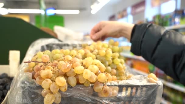 The hand of a woman wearing a down jacket selects a bunch of grapes in a supermarket - Video