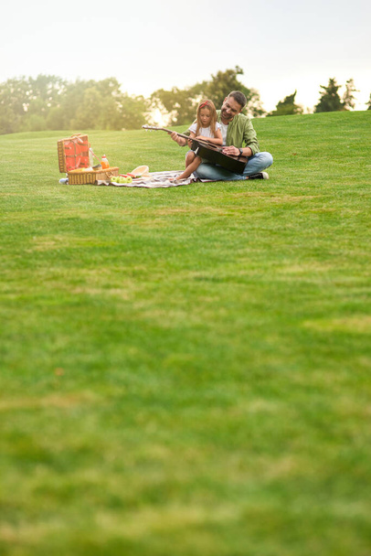 Plan complet de la petite fille heureuse assise avec son père aimant sur une herbe verte dans le parc et apprenant à jouer de la guitare - Photo, image