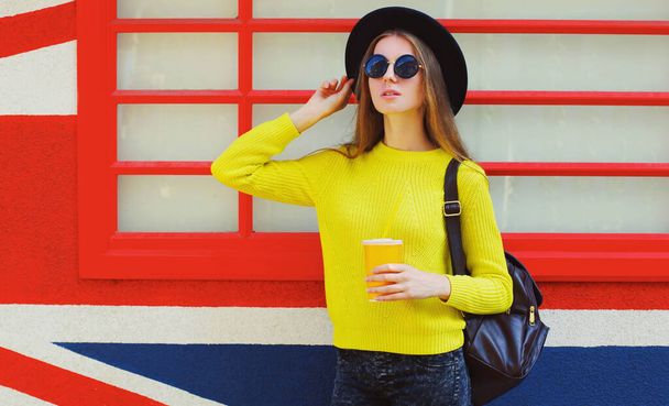 Portrait of stylish young woman with cup of juice wearing a yellow sweater, black round hat over background - Φωτογραφία, εικόνα
