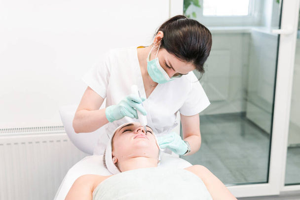 O cosmetologist feminino faz um procedimento da cara de limpeza ultrasônica, close-up. Uma jovem num salão de beleza. Conceito de cosmetologia e cuidados com a pele profissional - Foto, Imagem