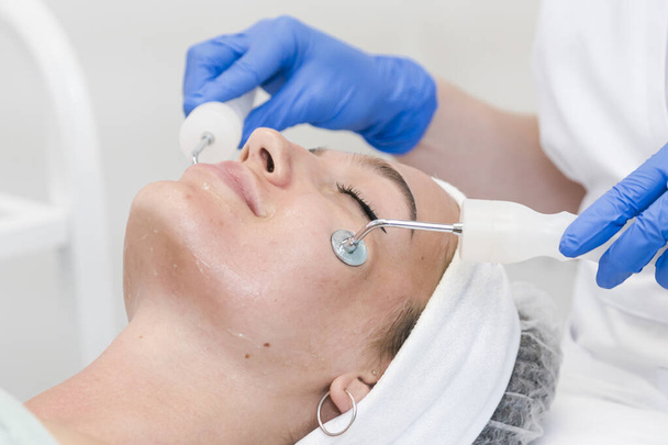 The young female client of cosmetic salon having microcurrent procedure on her face with special devices, close-up. Beautician using electrical impulses for facial procedures. Concepts of skin care product and beauty salon or clinic. - Foto, Bild