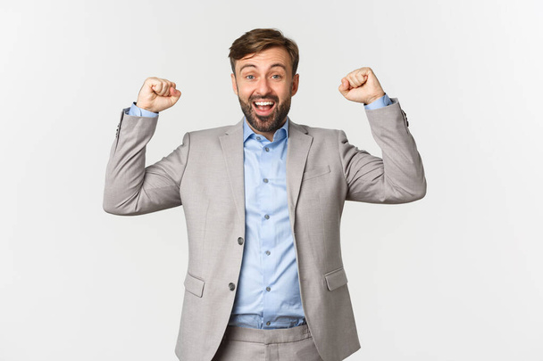 Портрет полегшеного і щасливого бородатого чоловіка в діловому костюмі, радіючи над перемогою, піднімаючи руки і кажучи так із задоволенням, стоячи над білим тлом
 - Фото, зображення