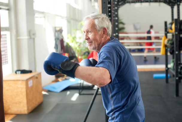 Hombre mayor supera saco de boxeo en gimnasio. - Foto, imagen