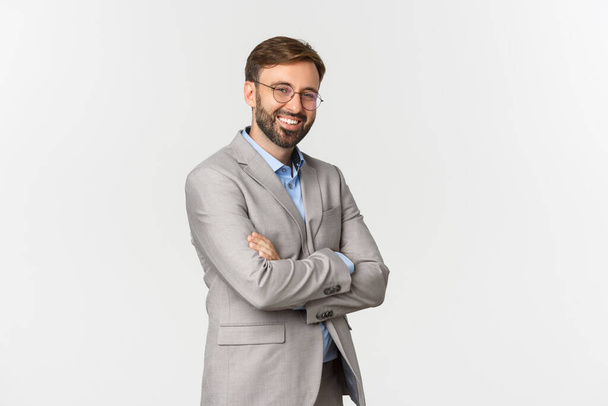 Retrato de empresário confiante com barba, vestindo terno cinza e óculos, braços cruzados no peito e sorrindo auto-confiante, em pé sobre fundo branco - Foto, Imagem