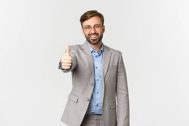 Retrato de hombre de negocios guapo sonriente con barba, con gafas y traje gris, mostrando los pulgares hacia arriba satisfecho, alabando el buen trabajo, de pie sobre fondo blanco - Foto, Imagen