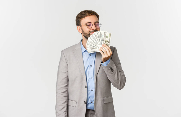 Portrait of successful happy businessman, wearing grey suit and glasses, smelling money and smiling delighted, standing over white background - Photo, Image