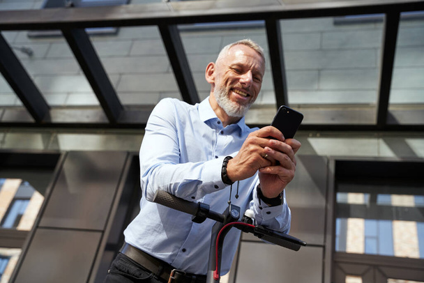 Generování nápadů. Veselý podnikatel středního věku s úsměvem při použití svého smartphonu, stojí s elektrickým skútrem venku v blízkosti moderní budovy - Fotografie, Obrázek
