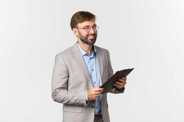 Zdjęcie profilowe przystojnego, pewnego siebie biznesmena, noszącego okulary i szary garnitur, trzymającego schowek i patrzącego w prawo, uśmiechającego się podczas prezentacji, białe tło - Zdjęcie, obraz