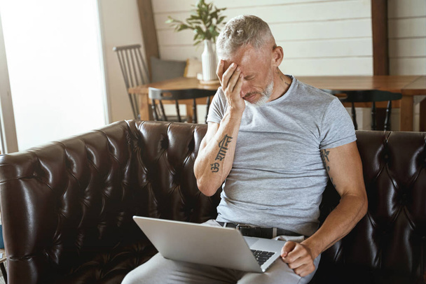 frustrovaný muž středního věku se dotýká hlavy a cítí stres, zatímco sedí s notebookem na pohovce v obývacím pokoji a pracuje na dálku - Fotografie, Obrázek