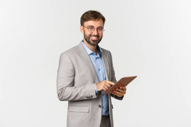 Ritratto di un bell'imprenditore barbuto in abito grigio e occhiali, con tablet digitale e lavoro, sorridente felice davanti alla macchina fotografica, in piedi su sfondo bianco - Foto, immagini