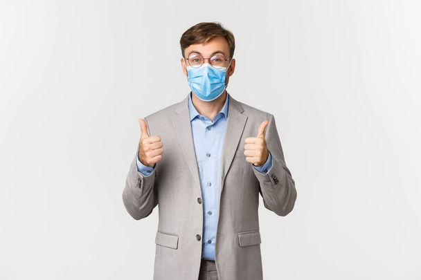 Concept of work, covid-19 and social distancing. Portrait of cheerful businessman in gray suit and medical mask, showing thumbs-up in approval, recommend something, white background - Photo, Image