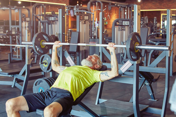 Тяжело работать. Сильный взрослый мужчина в спортивной одежде, поднимающий штангу или штангу в спортзале - Фото, изображение