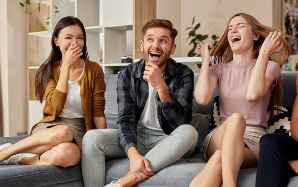 Risas contagiosas. Grupo de alegres jóvenes amigos multiculturales en ropa casual disfrutando del tiempo juntos mientras se sientan en el sofá en el apartamento moderno - Foto, Imagen