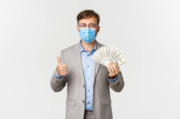 Concept of business, covid-19 and social distancing. Portrait of businessman in medical mask, showing salary money and thumbs-up, standing over white background - Φωτογραφία, εικόνα