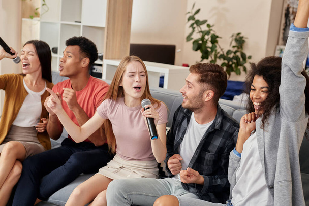 Amigos e música. Amigos alegres olhando animado ao jogar karaoke em casa, cantando com microfone, sentado no sofá na sala de estar - Foto, Imagem