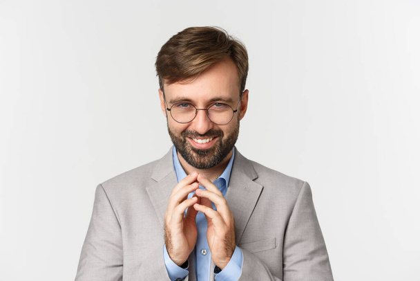 Close-up of bearded businessman in glasses and suit, scheming something, smiling devious and having perfect plan, standing over white background - Foto, Imagem