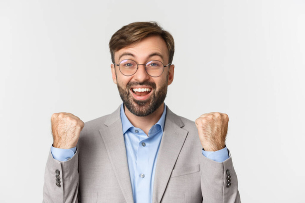 Close-up of successful businessman in glasses and gray suit, rejoicing, saying yes with happy face and making fist pump, achieve goal, standing over white background - Φωτογραφία, εικόνα