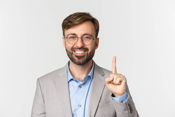 Close-up of handsome bearded man in glasses and gray suit, showing number one and smiling, standing over white background - Foto, Imagen