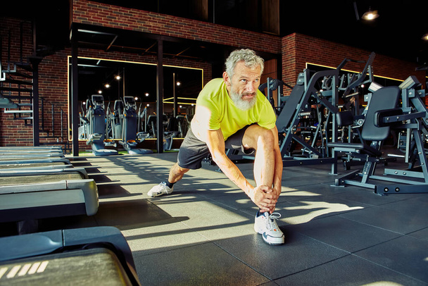 Warming up. Portrait of athletic middle aged man in sportswear stretching legs before training in a gym or sport club, full length - Photo, image