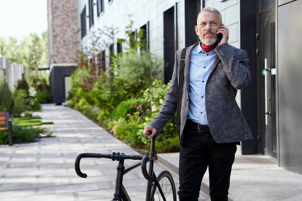 Κομψός ώριμος επιχειρηματίας κοιτάζει αλλού ενώ μιλάει στο τηλέφωνο, στέκεται με το ποδήλατο έξω σε μια μέρα - Φωτογραφία, εικόνα