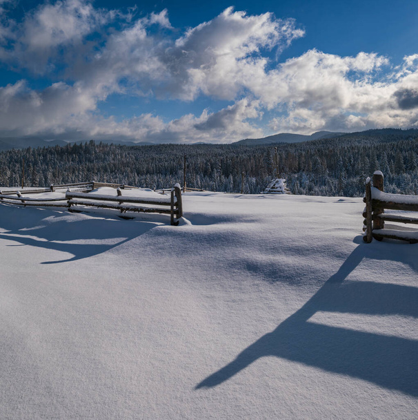 Picturesque shadows on snow from wood fence. Alpine mountain winter hamlet outskirts, snowy path, fir forest on far misty and cloudy hills. - Fotoğraf, Görsel