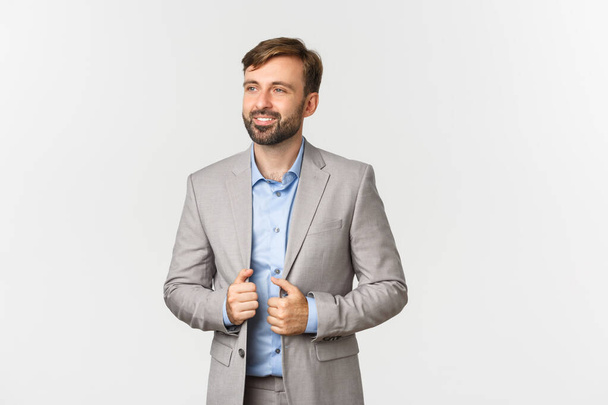 Imagem de empresário bem sucedido e confiante em terno cinza e camisa azul, sorrindo satisfeito e olhando para a esquerda, em pé sobre fundo branco - Foto, Imagem
